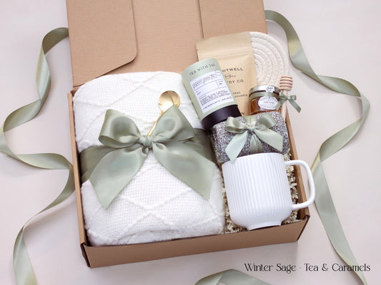 A soft-toned gift box with a white textured blanket, white mug, honey jar, tea tube, caramel packet, and cozy socks tied in sage ribbon.