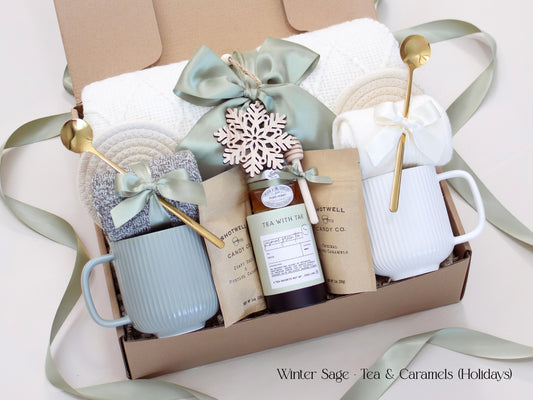 A festive Winter Sage holiday gift box for couples with a white knit blanket, two mugs, cozy socks, artisan tea, gourmet caramels, and a wooden snowflake ornament. Finished with soft sage ribbons, perfect for holiday gifting.