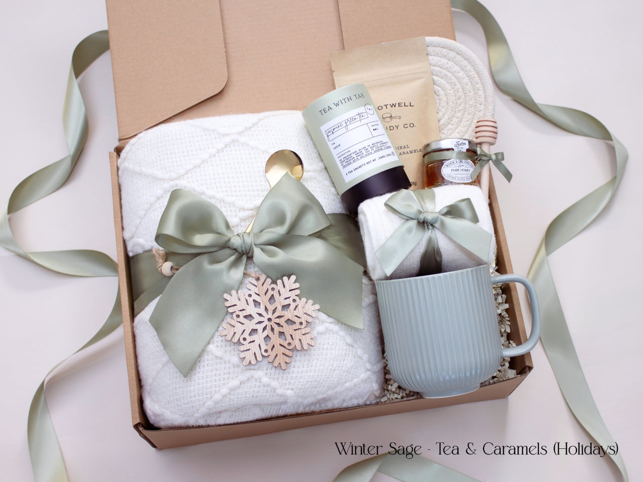 A festive winter gift box with a white blanket, mug, honey, tea, caramels, and socks tied with sage ribbon, adorned with a wooden snowflake ornament for a holiday touch.