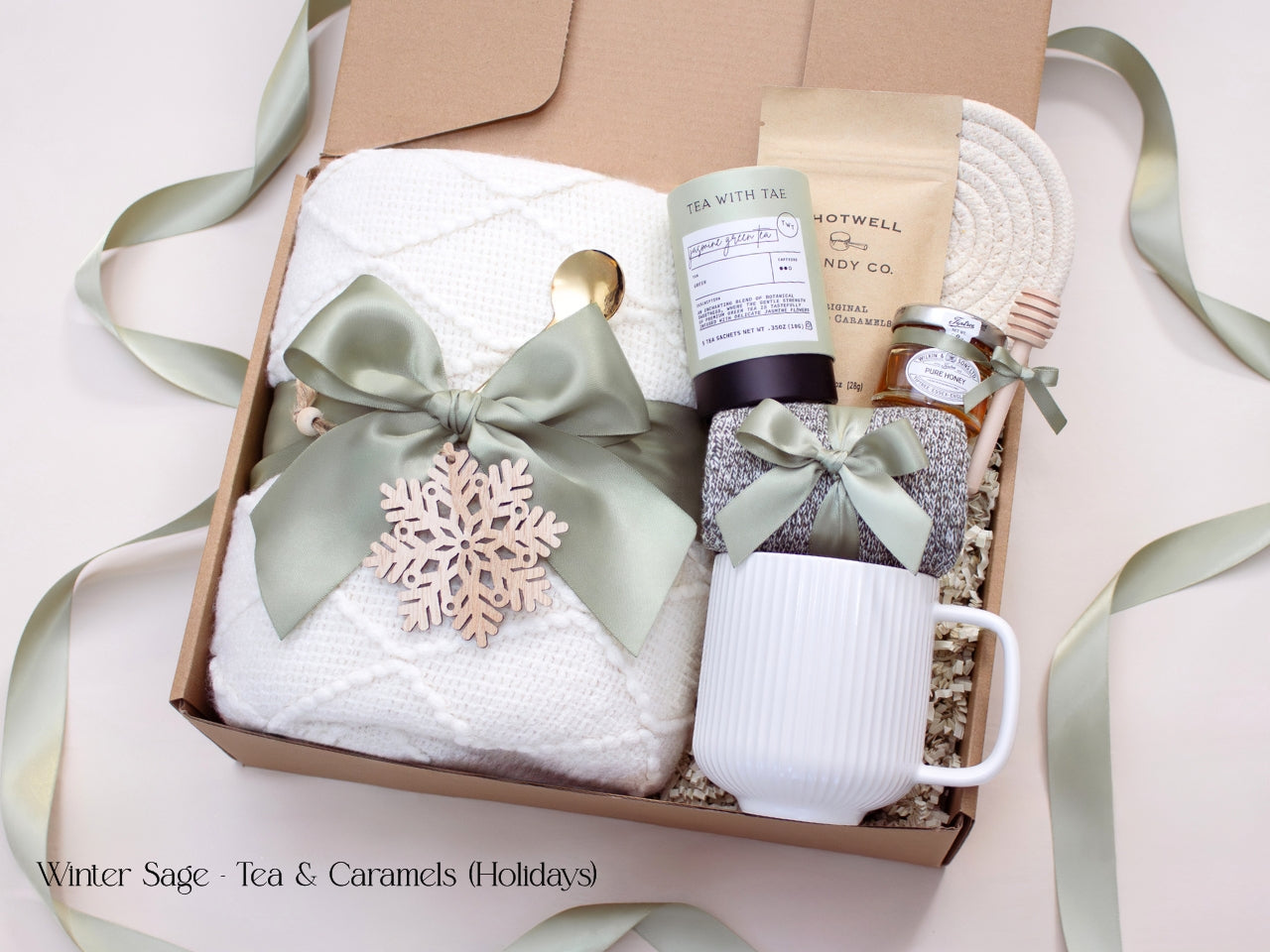 A festive winter gift box with a white blanket, mug, honey, tea, caramels, and socks tied with sage ribbon, adorned with a wooden snowflake ornament for a holiday touch.