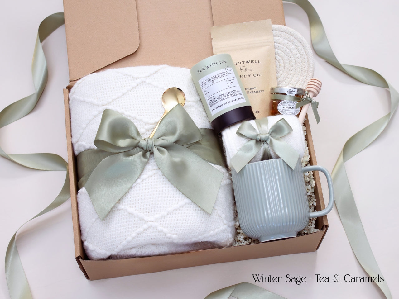 A soft-toned gift box with a white textured blanket, white mug, honey jar, tea tube, caramel packet, and cozy socks tied in sage ribbon.
