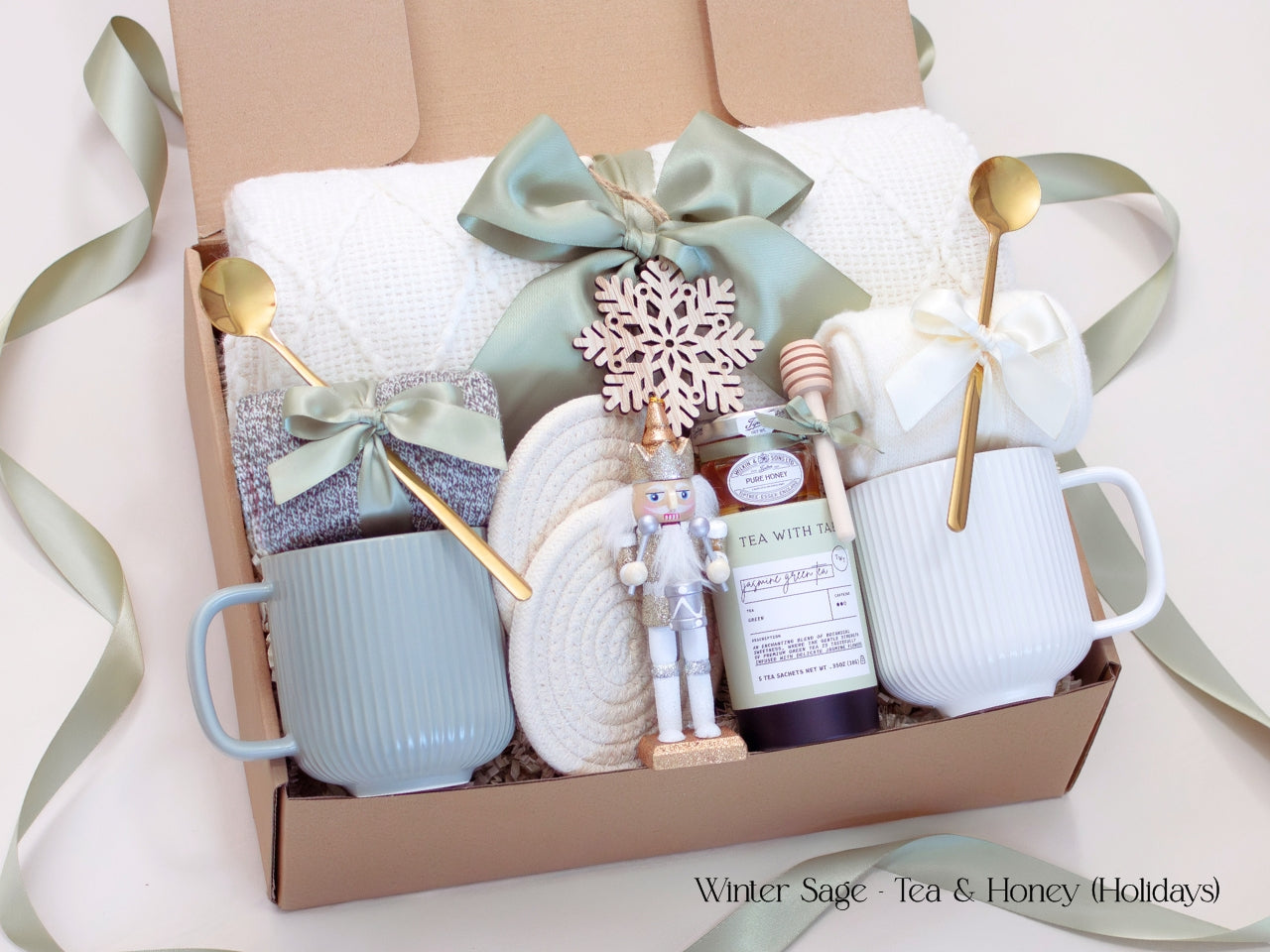A luxurious Winter Sage holiday gift box for couples featuring a white knit blanket, two mugs, artisan tea, pure honey, cotton coasters, a wooden nutcraker and a wooden snowflake ornament. Styled in calming sage and ivory tones with satin ribbon.