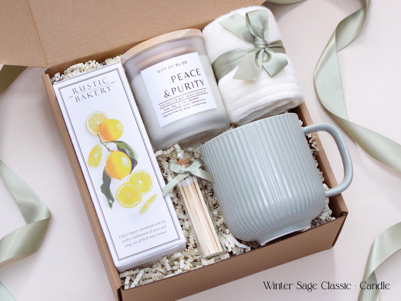 Neutral-toned gift box with a white candle, ribbed sage mug, shortbread cookies, cozy socks, and matches tied with a satin sage ribbon, exuding calm and minimal winter elegance.