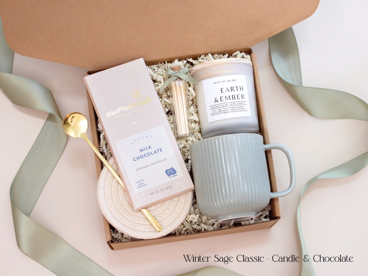 A minimalist winter gift box featuring a Moonstruck chocolate bar, Earth & Ember candle, ribbed sage mug, gold spoon, and cotton rope coasters, styled with sage green ribbon for a refined, cozy look.