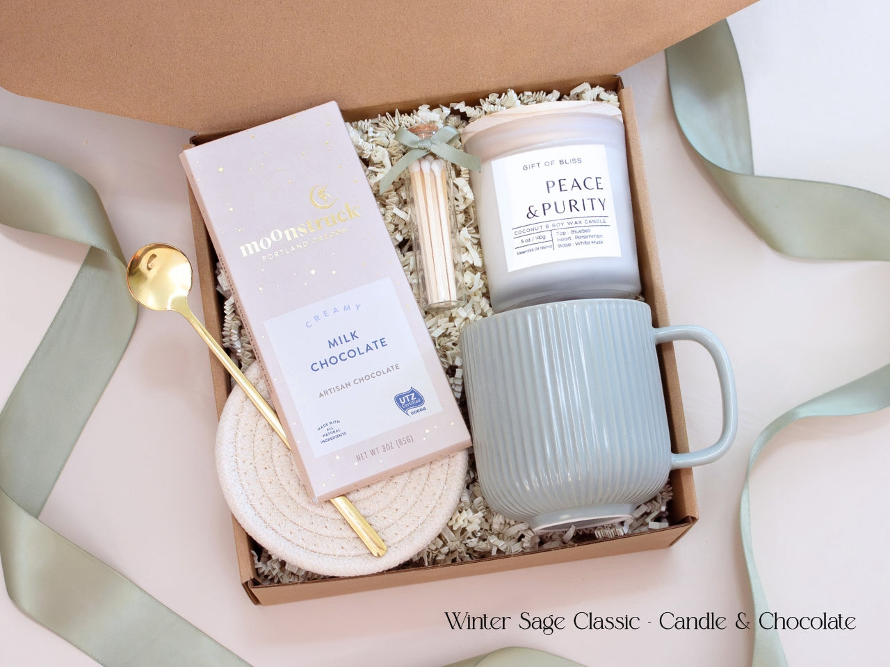 A minimalist winter gift box featuring a Moonstruck chocolate bar, Earth & Ember candle, ribbed sage mug, gold spoon, and cotton rope coasters, styled with sage green ribbon for a refined, cozy look.