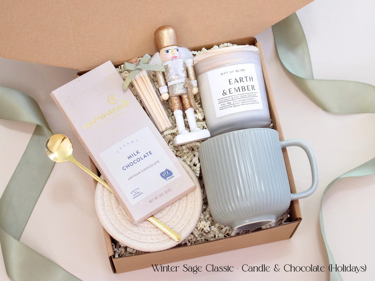 Holiday-themed neutral gift box with a Moonstruck chocolate bar, Earth & Ember candle, sage mug, gold spoon, and nutcracker figurine, styled with sage ribbon and shredded ivory filler for a modern festive touch.
