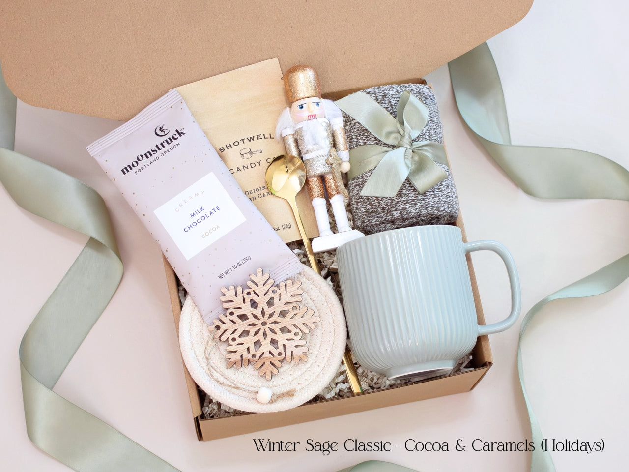 A festive gift box featuring a Moonstruck cocoa packet, caramel candies, nutcracker figurine, sage mug, and wooden snowflake ornament, tied with sage ribbon for a refined winter presentation.