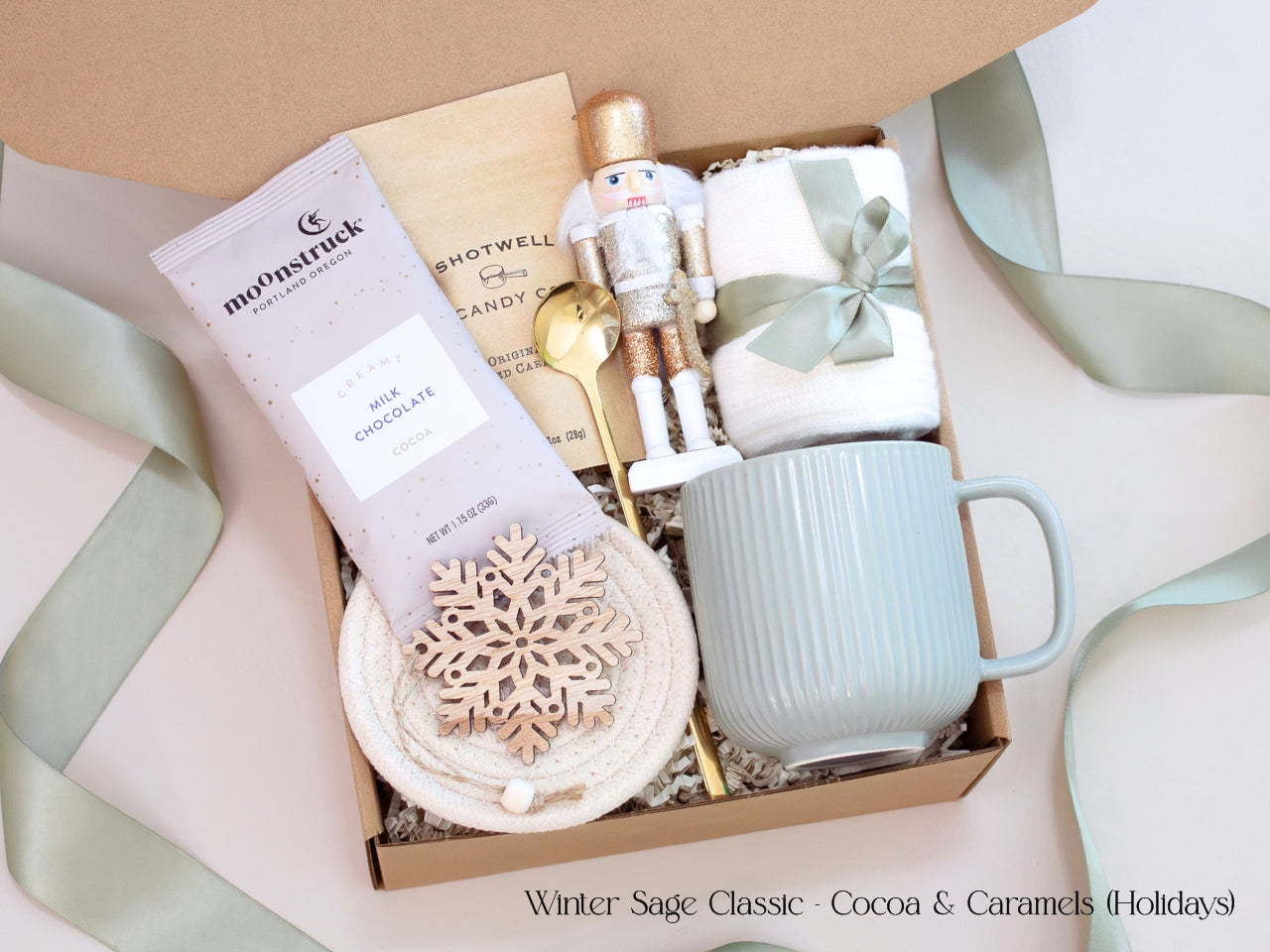 A festive gift box featuring a Moonstruck cocoa packet, caramel candies, nutcracker figurine, sage mug, and wooden snowflake ornament, tied with sage ribbon for a refined winter presentation.