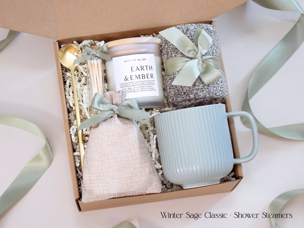 A neutral winter self-care gift box featuring a ribbed sage mug, candle, cozy socks, gold spoon, and linen pouch of shower steamers tied with sage ribbon.