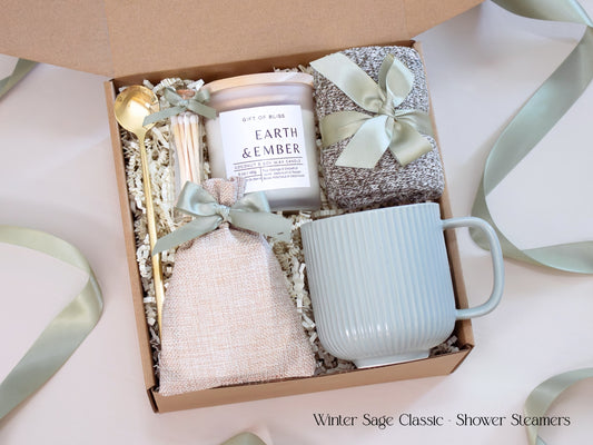 A neutral winter self-care gift box featuring a ribbed sage mug, candle, cozy socks, gold spoon, and linen pouch of shower steamers tied with sage ribbon.