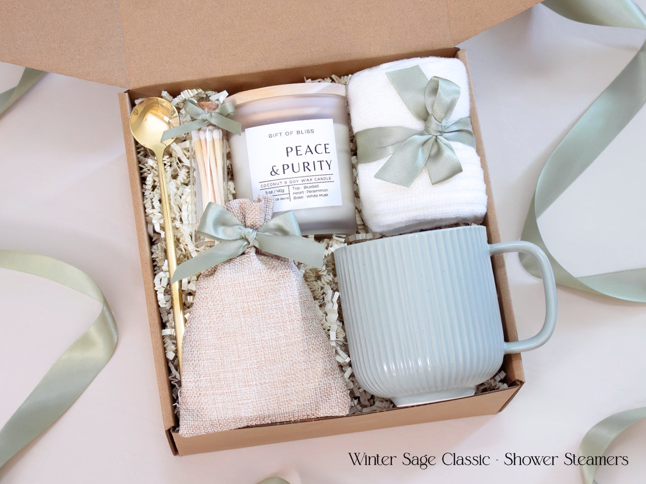 A neutral winter self-care gift box featuring a ribbed sage mug, candle, cozy socks, gold spoon, and linen pouch of shower steamers tied with sage ribbon.