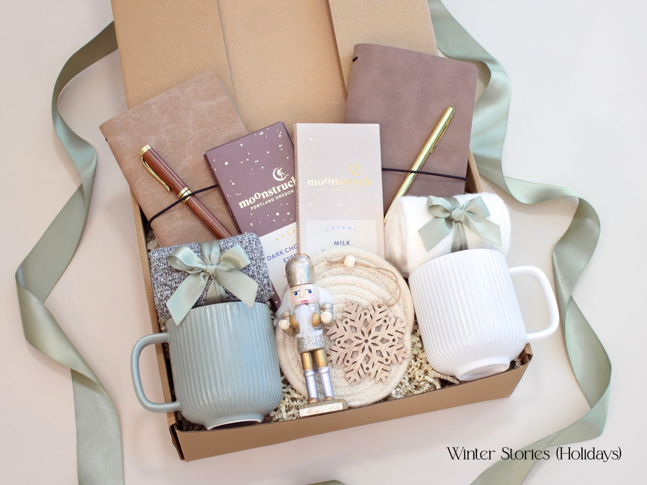 A holiday-themed Winter Stories gift box for couples featuring two mugs, journals with pens, cozy socks, Moonstruck chocolates, a nutcracker accent, and a wooden snowflake ornament. Wrapped with a soft sage ribbon, perfect for festive gifting.