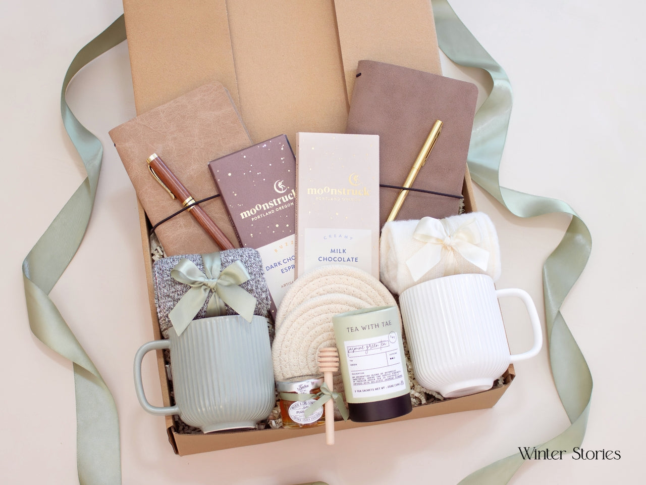 An elegant Winter Stories gift box for couples including two mugs, soft socks, Tea with Tae tea, Moonstruck chocolates, journals with pens, and a honey jar. Presented with a sage ribbon for a cozy and thoughtful gift.